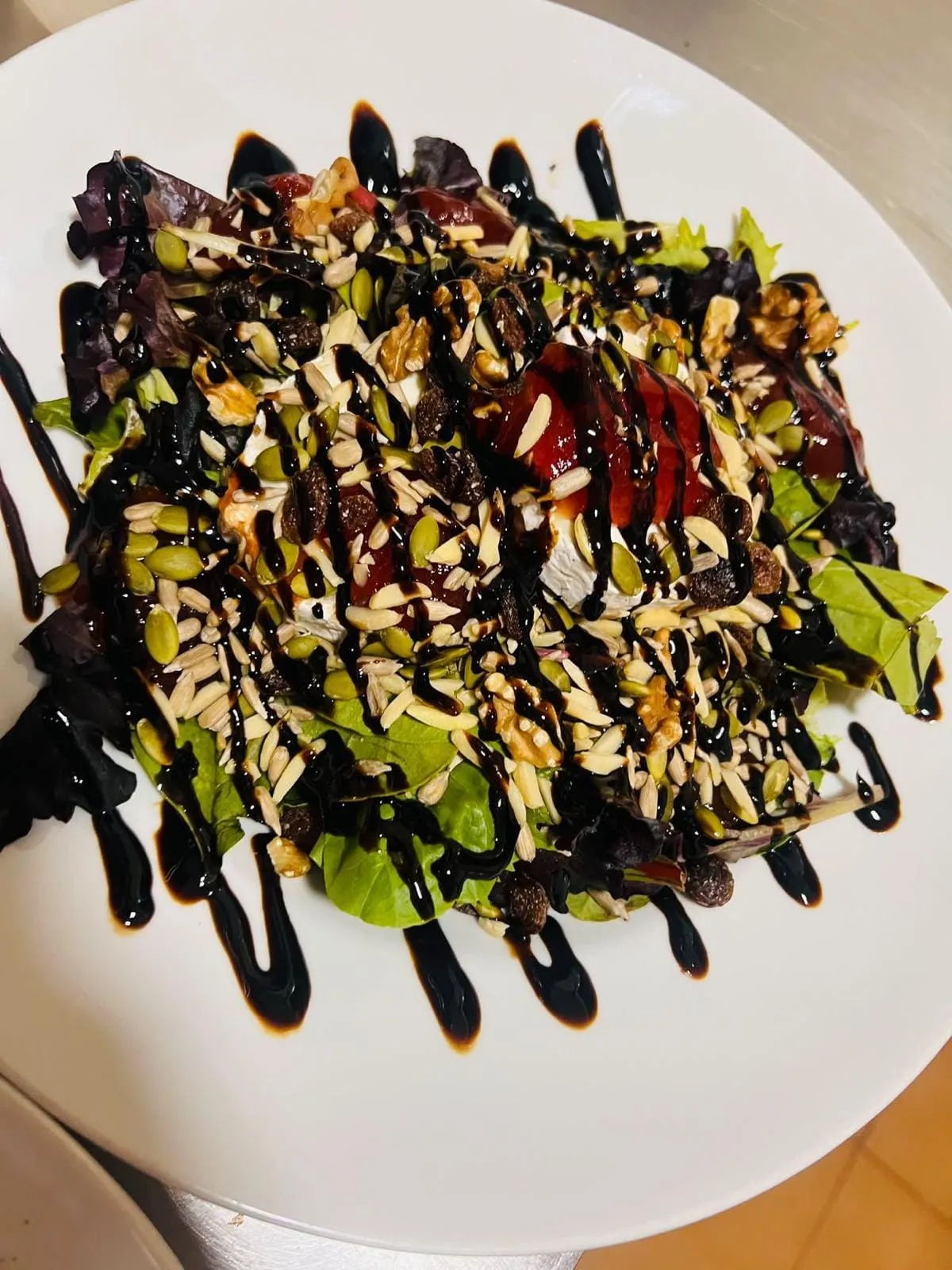 Ensalada de brotes con rodajas de queso de cabra cubiertas de mermelada roja, nueces, almendra laminada, pipas y pasas, con reducción de balsámico por encima.