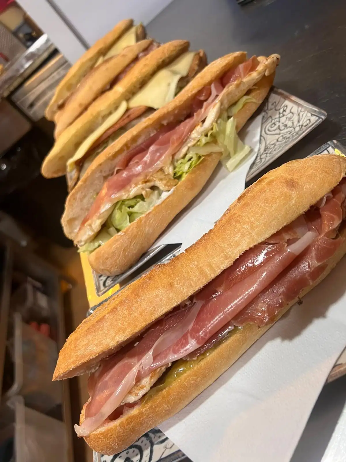 Tres bocadillos de barra sobre servilletas: uno de jamón serrano con huevo, otro con lechuga y carne a la plancha, y otro con queso fundido.