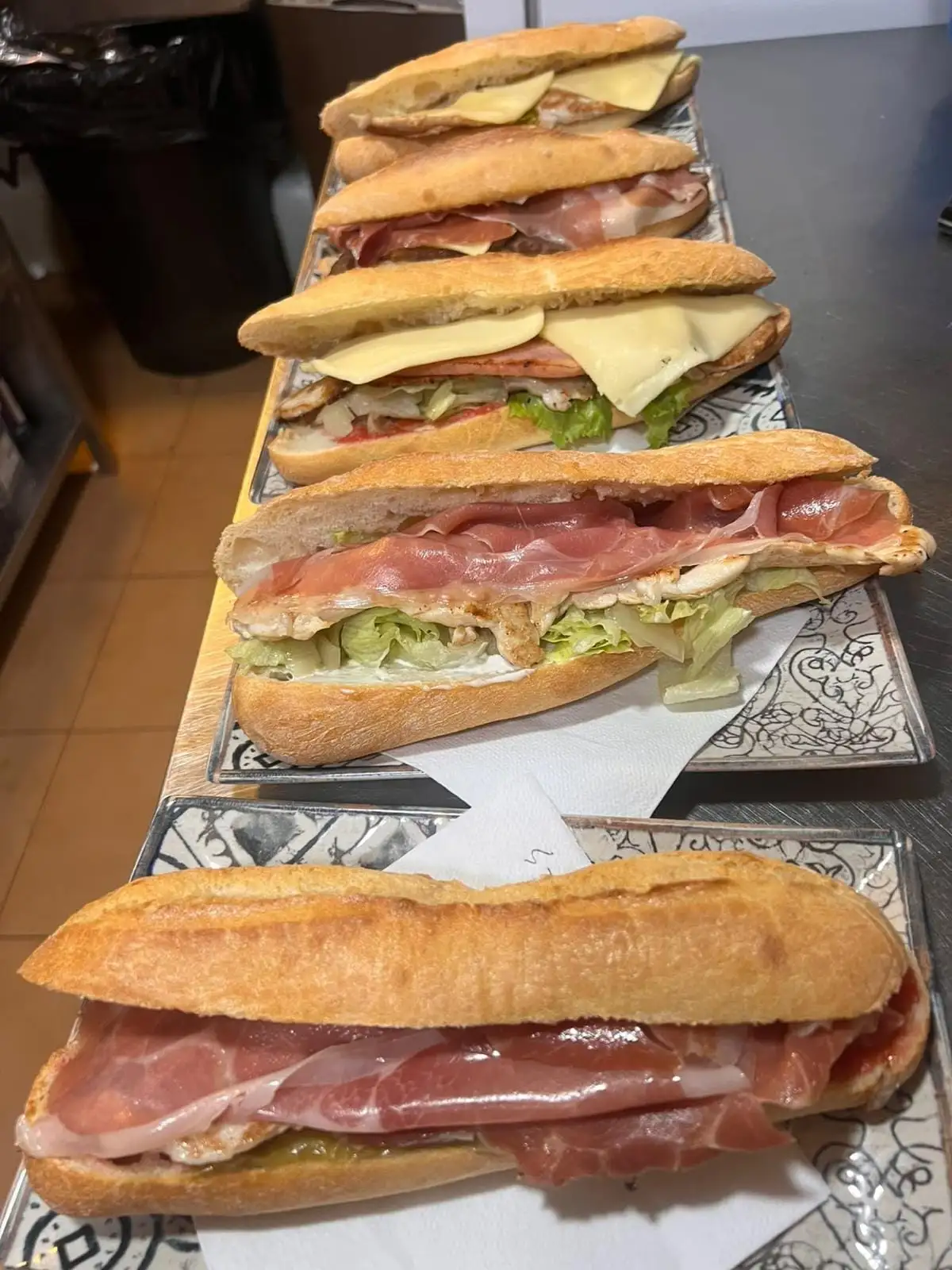 Cinco bocadillos de barra en fila sobre bandejas: jamón serrano, carne a la plancha con lechuga y queso fundido.