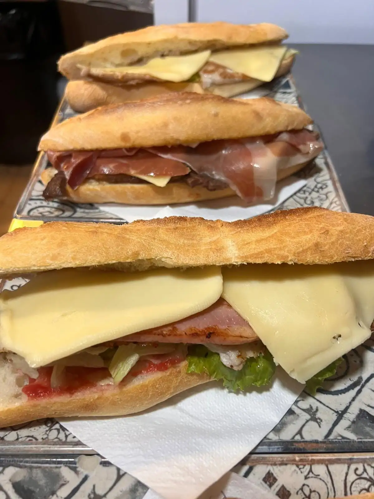 Bocadillo de barra con lonchas de queso, carne a la plancha, lechuga y tomate, con otros dos detrás en la misma barra.