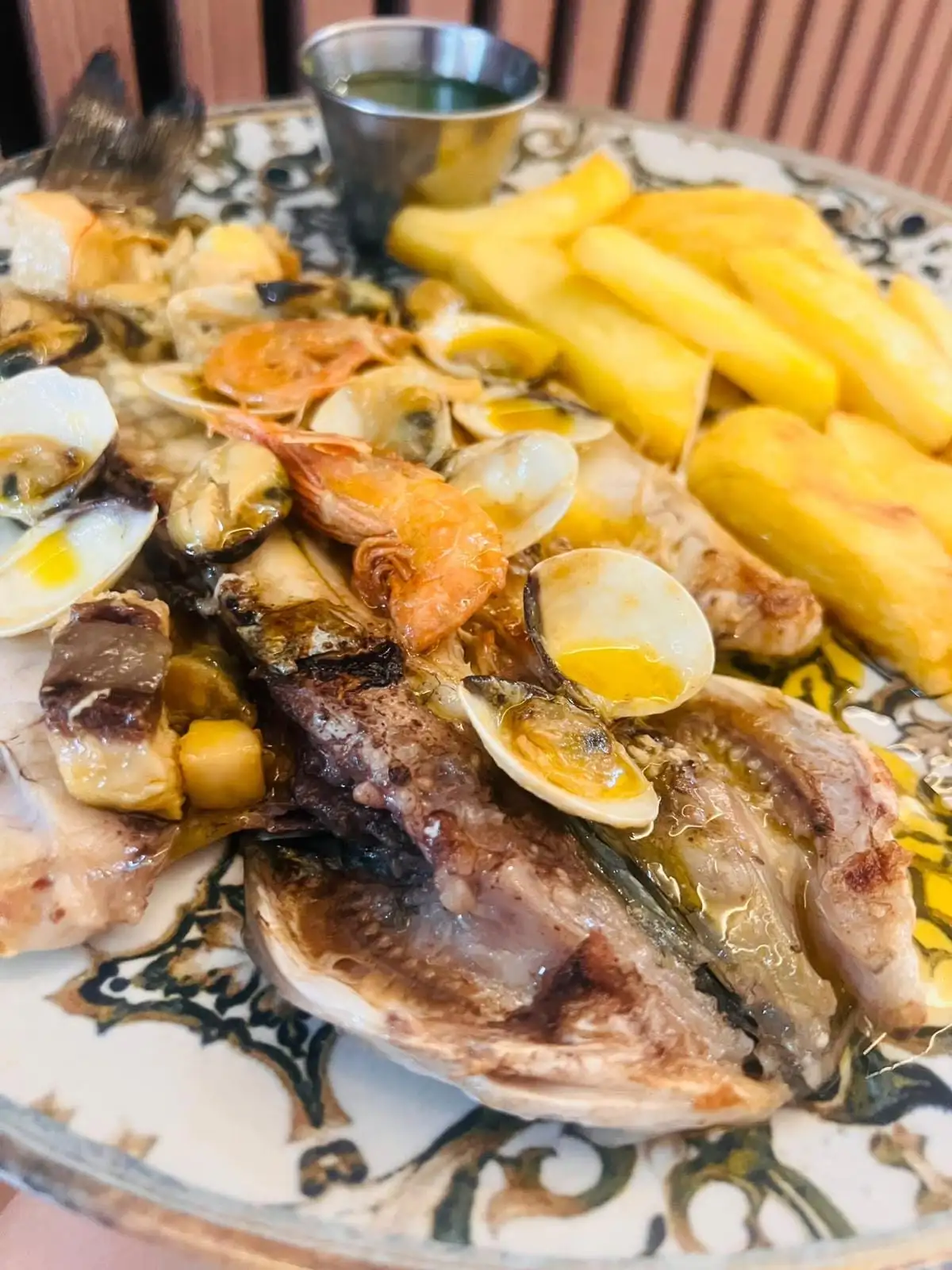 Pescado abierto a la plancha con gambas, almejas y mejillones, patatas fritas y un cuenco de salsa verde, en un plato decorado.