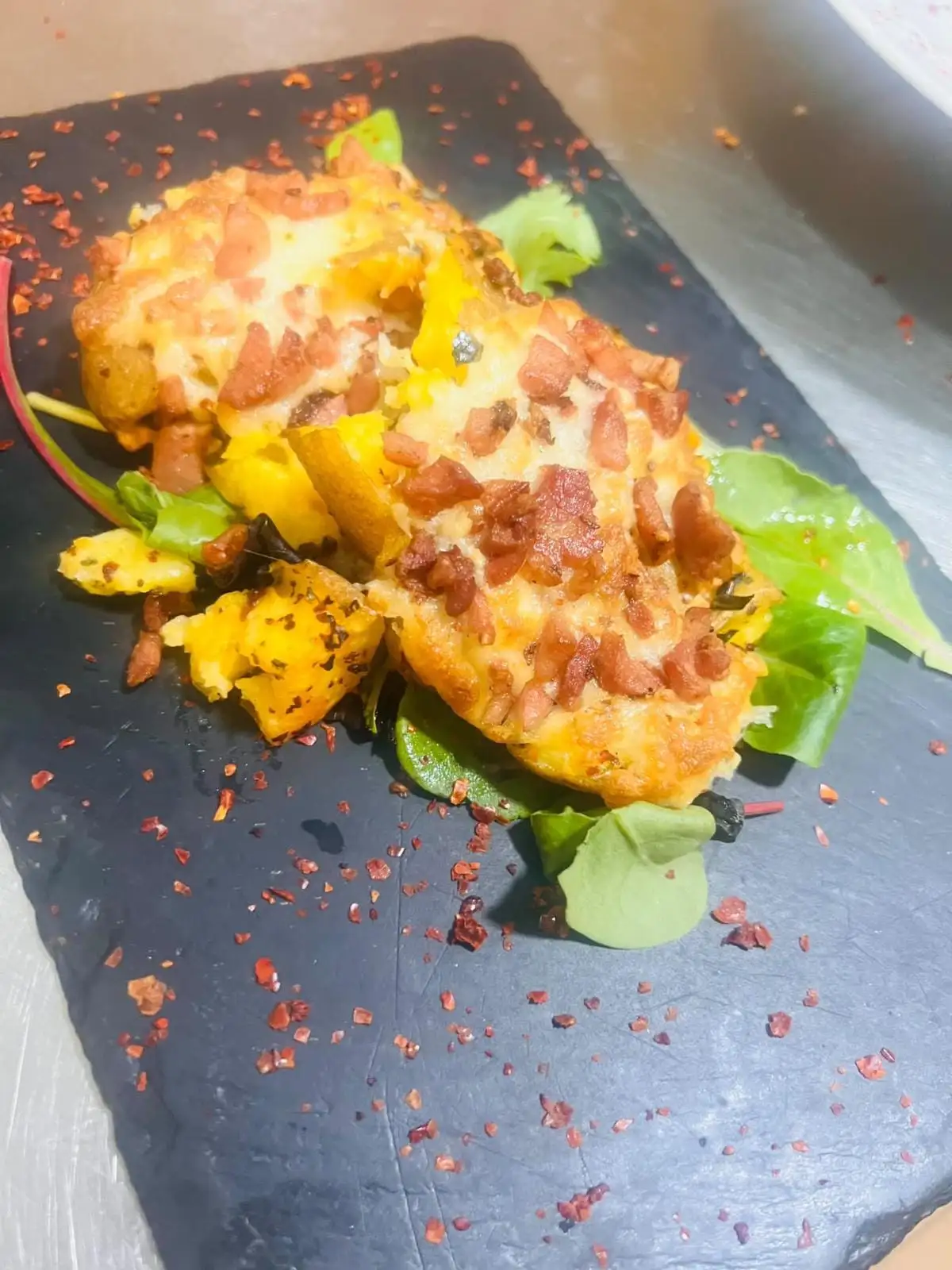 Patatas al horno gratinadas con queso fundido y taquitos de bacon, sobre pizarra con brotes verdes y copos de pimiento rojo.