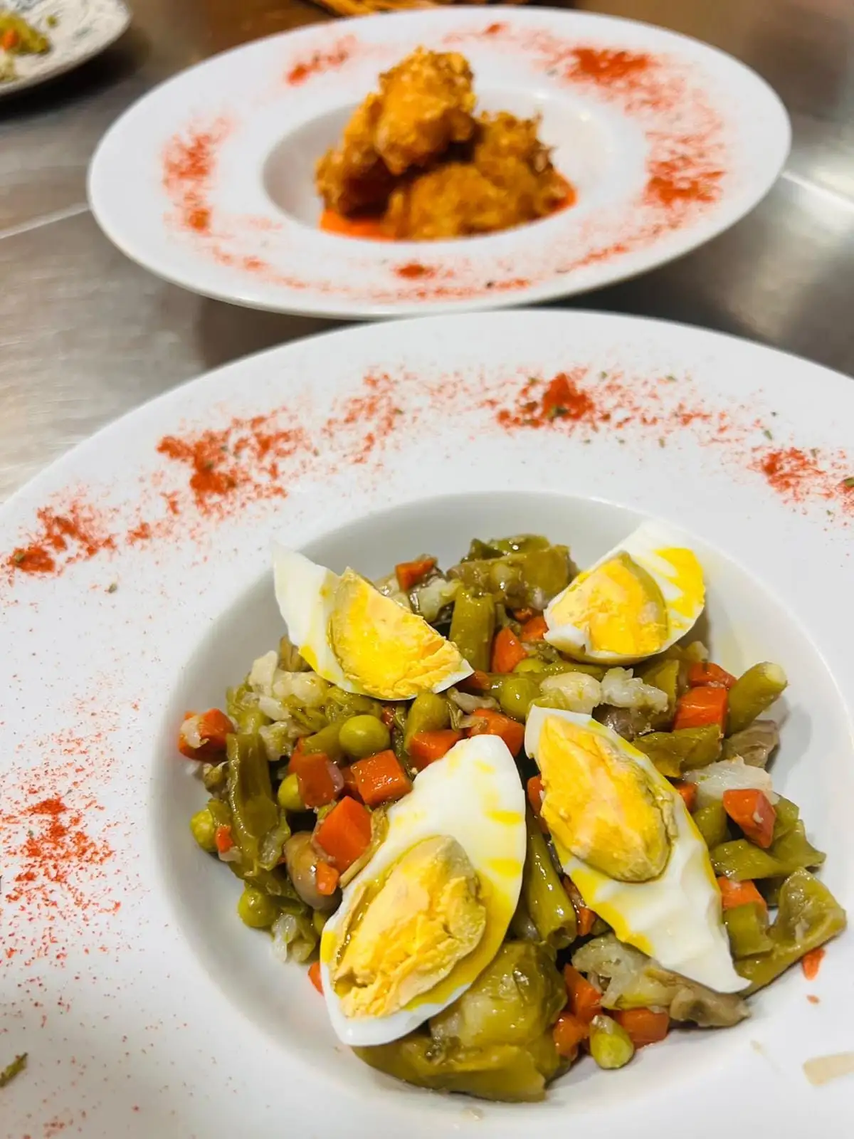 Menestra de verduras con judía verde, zanahoria, guisantes y alcachofa, coronada con cuartos de huevo duro y pimentón en el borde del plato.