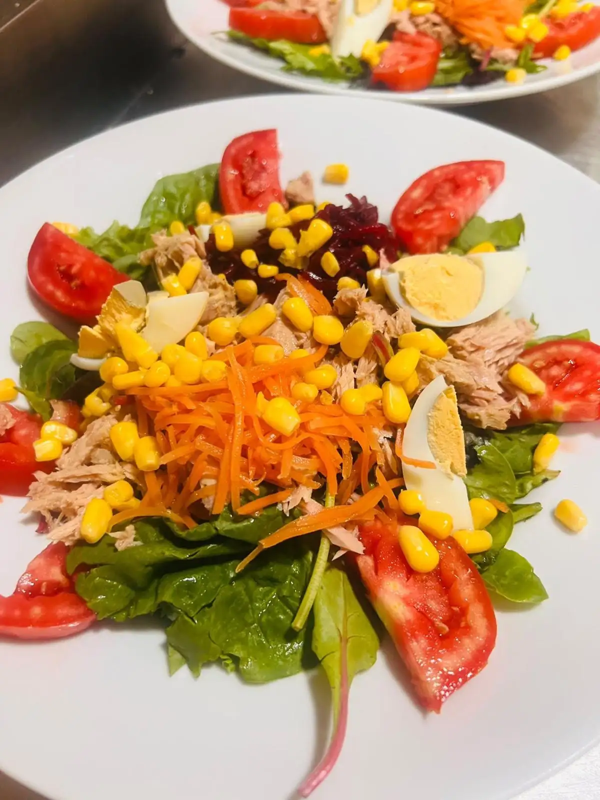 Ensalada mixta con lechuga, gajos de tomate, zanahoria y remolacha ralladas, maíz, huevo duro y atún, con otro plato igual al fondo.