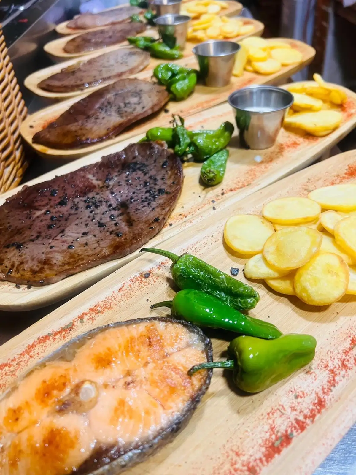 Hilera de tablas de madera con una rodaja de salmón a la plancha, filetes de carne, pimientos de Padrón, patatas en rodajas y salseras metálicas.