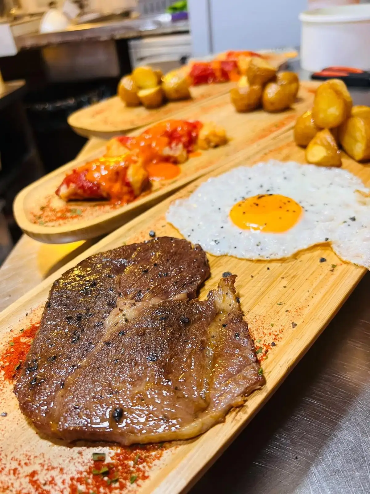 Tabla de madera con un filete a la plancha y un huevo frito, y al fondo otras tablas con patatas asadas y salsa roja.