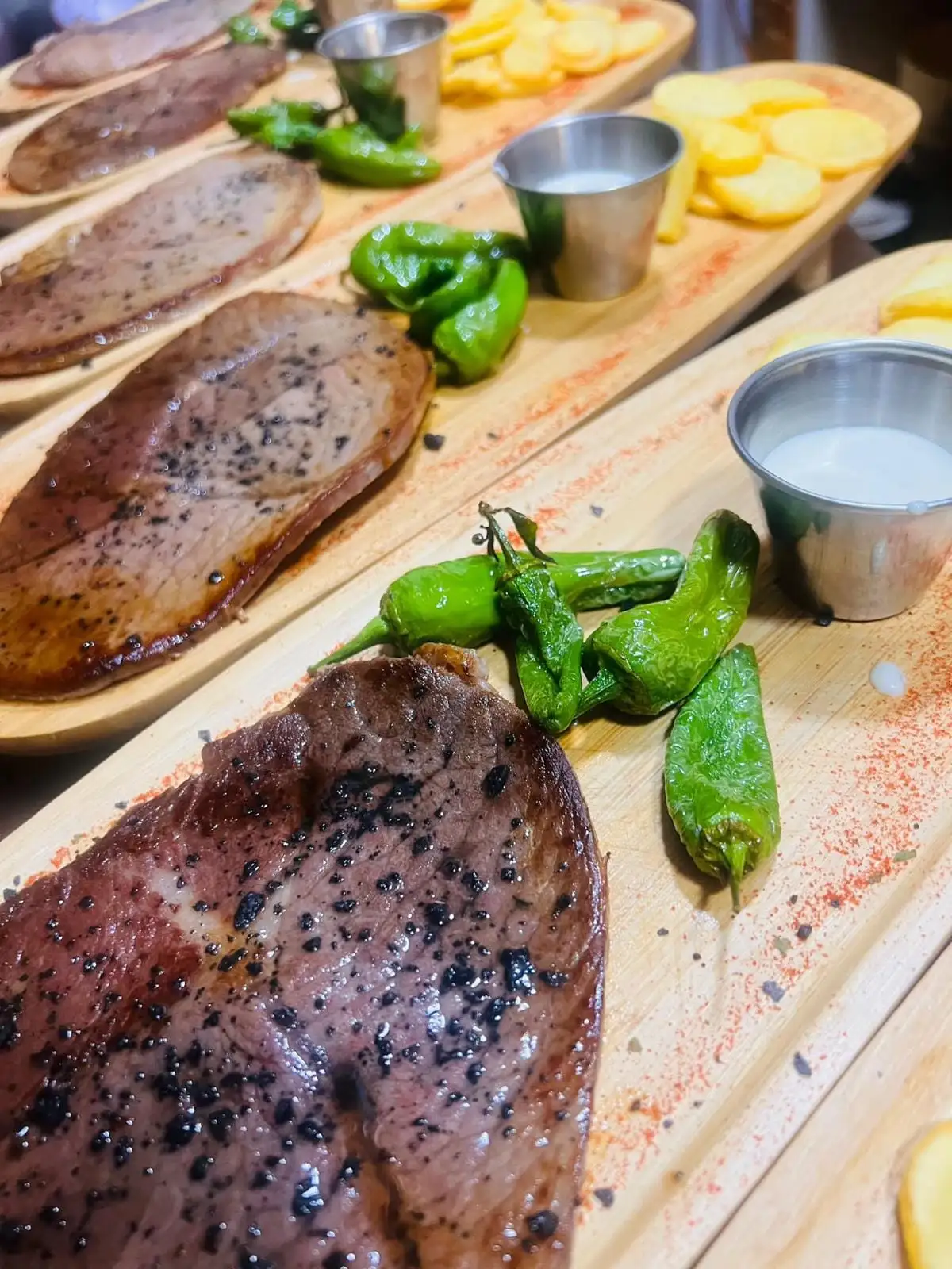 Hilera de tablas con filetes de carne a la plancha, pimientos de Padrón, salseras con salsa blanca y patatas en rodajas.