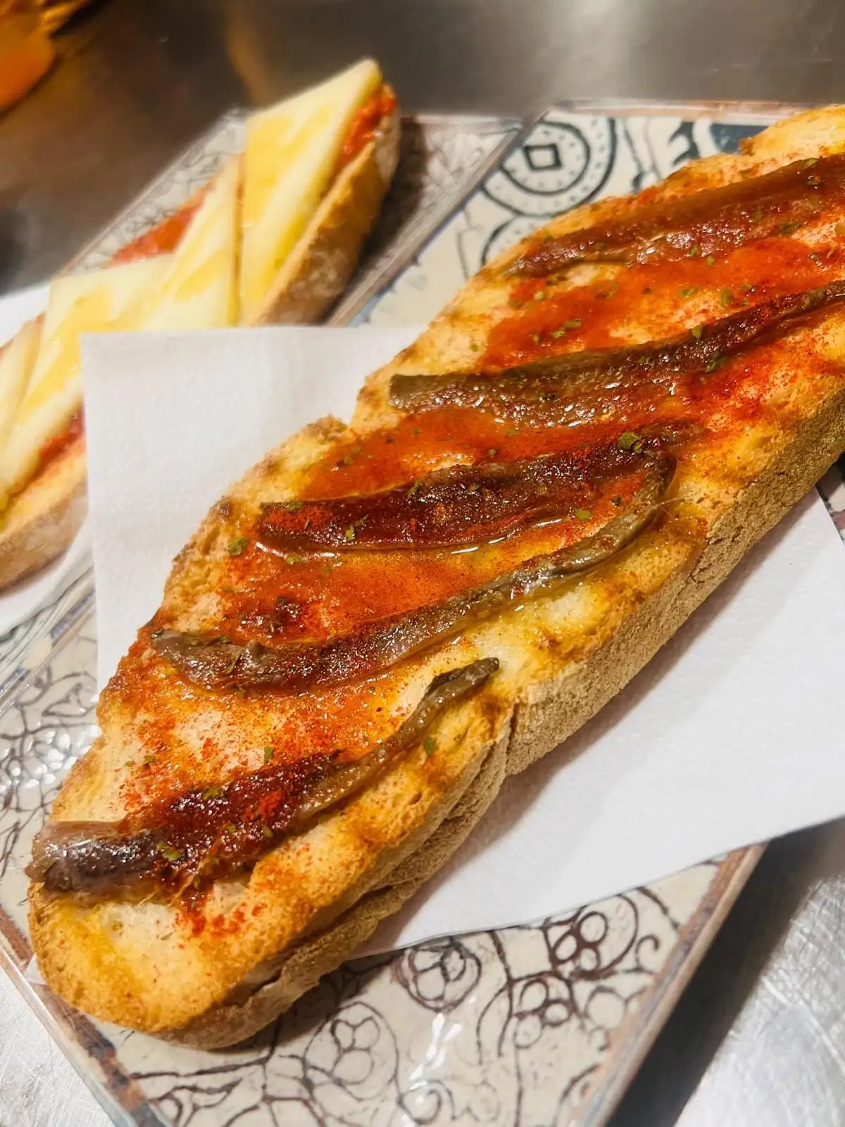 Media barra de pan tostado con tomate, anchoas y pimentón, y al fondo otra con tomate y queso en lonchas.