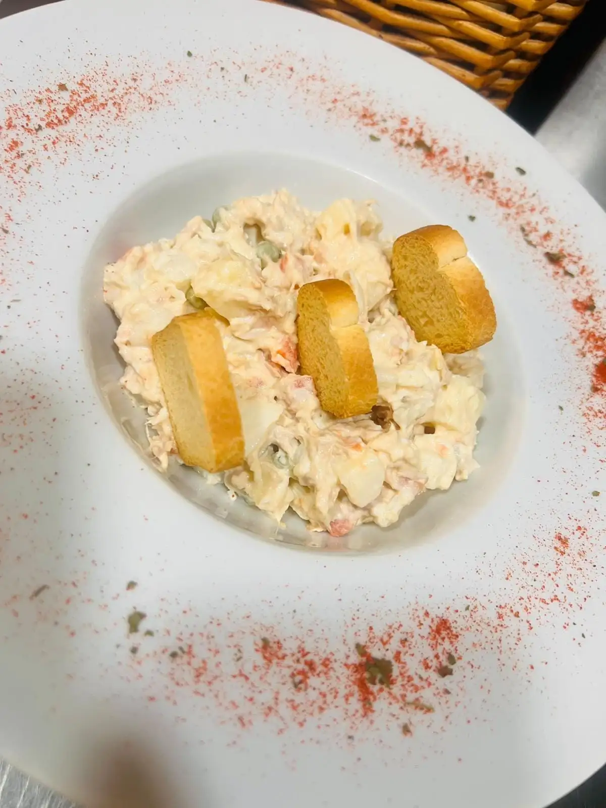 Ensaladilla con mayonesa, zanahoria y guisantes en un plato hondo, con tres rebanaditas de pan tostado encima.