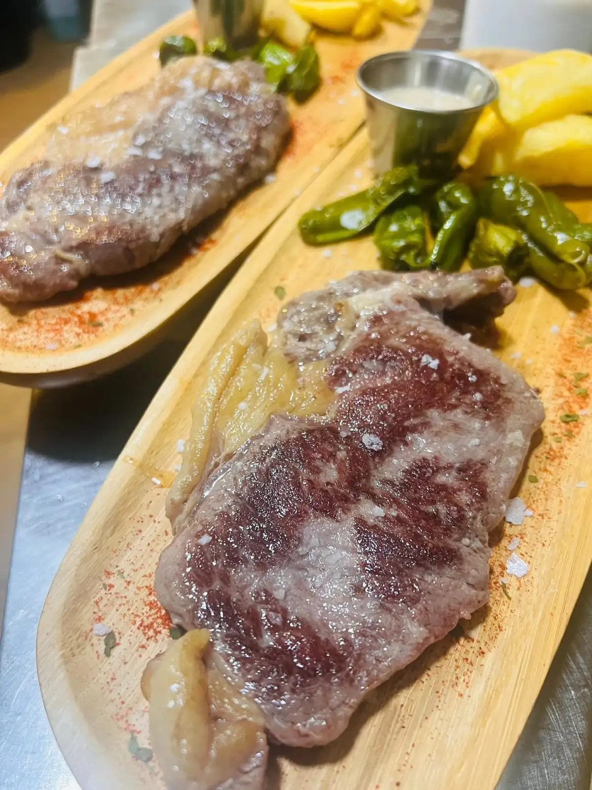 Dos entrecots a la plancha con sal gruesa sobre tablas de madera, con pimientos de Padrón, patatas fritas y una salsera con salsa blanca.