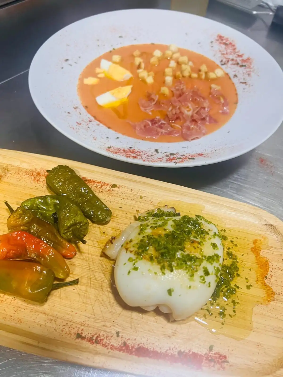 Tabla con sepia a la plancha con ajo y perejil y pimientos asados, y al fondo un plato de crema fría de tomate con huevo duro, picatostes y jamón.