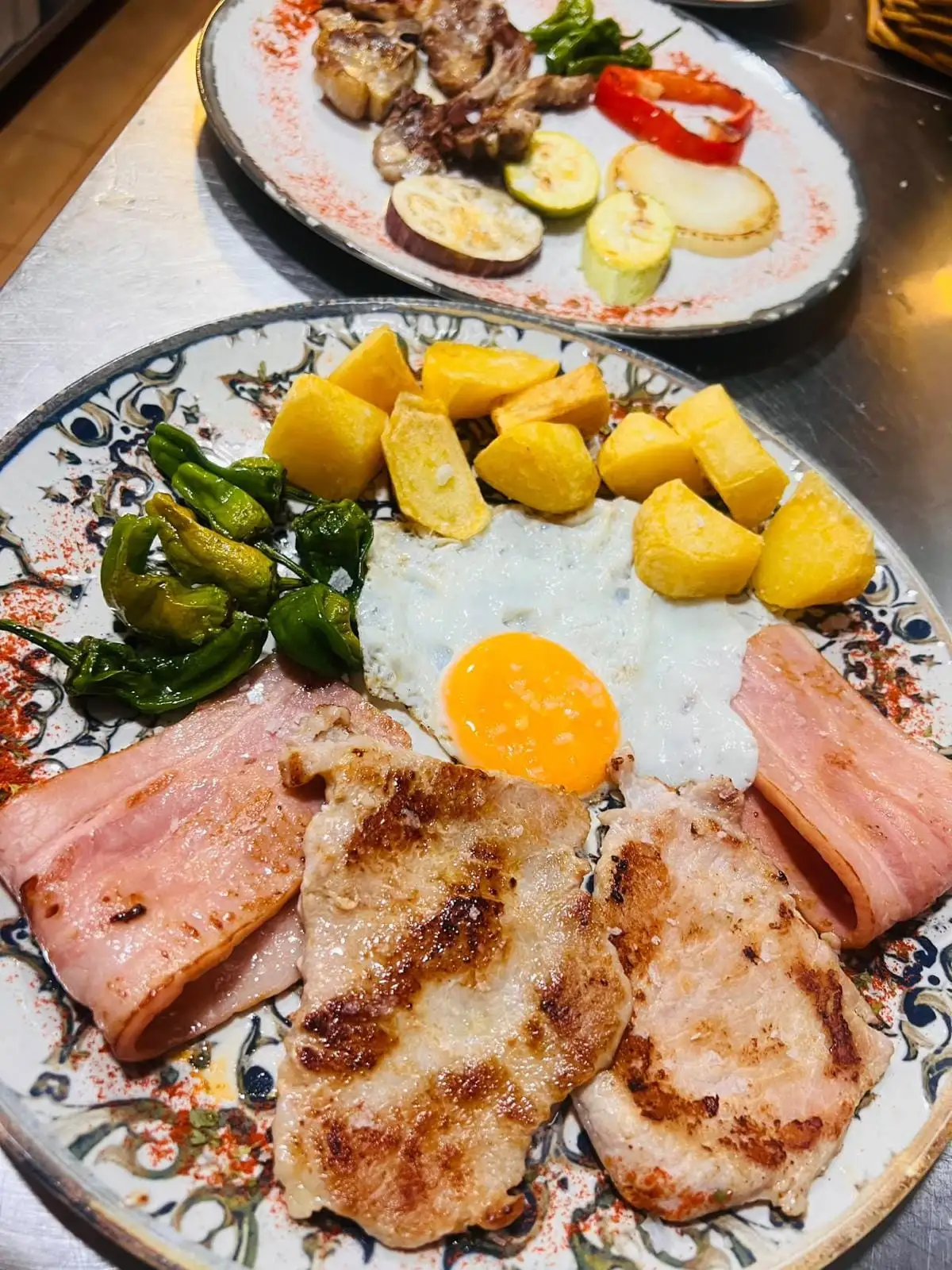 Plato con filetes a la plancha, lonchas de jamón york, huevo frito, patatas fritas en tacos y pimientos de Padrón, y al fondo otro plato con carne y verduras a la plancha.