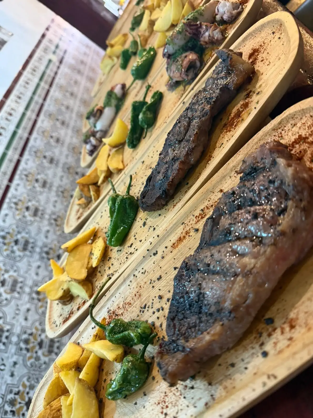 Hilera de tablas con filetes a la plancha, pimientos de Padrón, patatas en gajos y, al fondo, calamares con salsa verde.