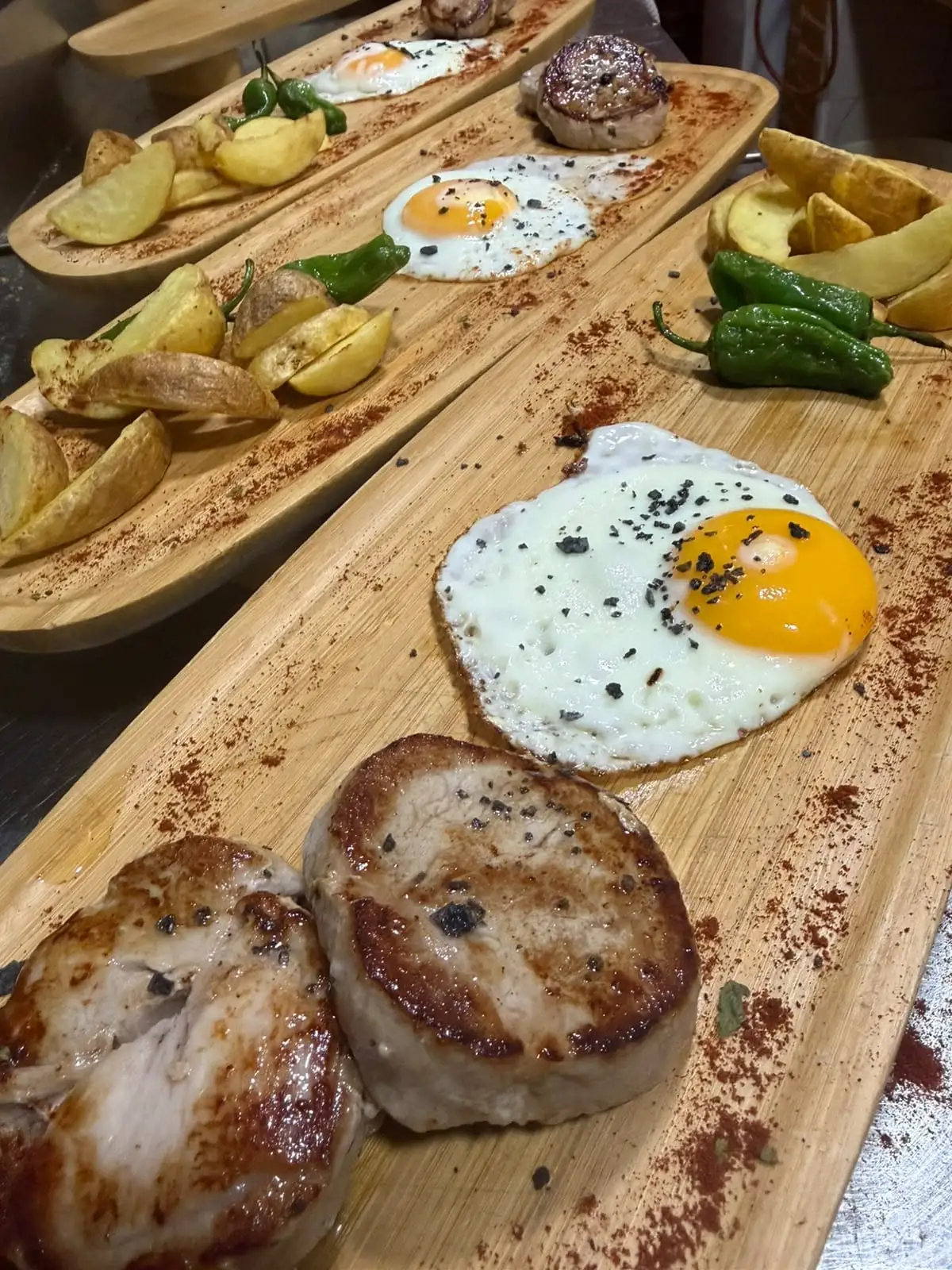 Tablas de madera alineadas, cada una con dos medallones de carne a la plancha, un huevo frito, patatas fritas en gajos y pimientos verdes.