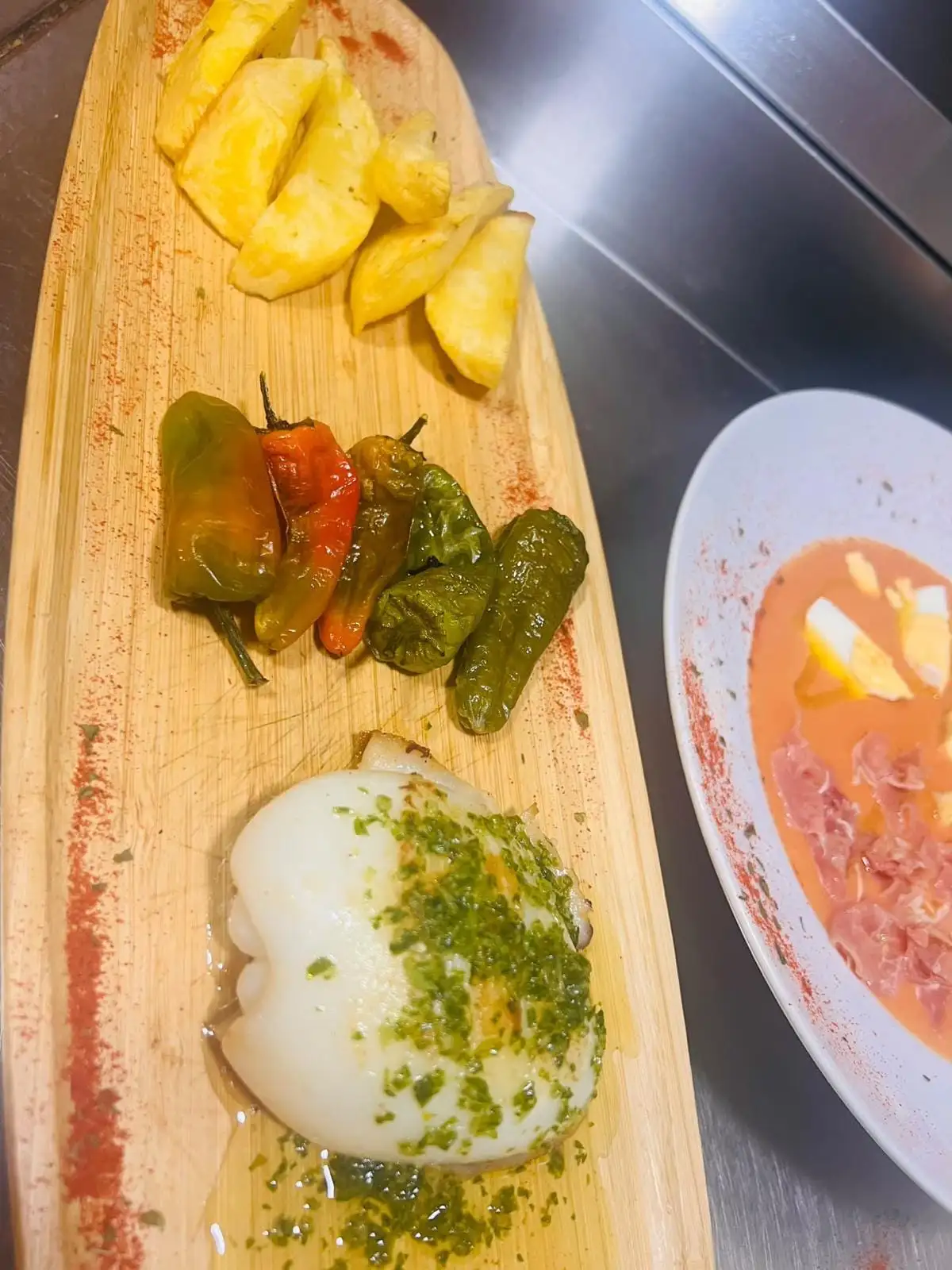 Tabla de madera con sepia a la plancha en salsa verde, pimientos de Padrón y patatas fritas, junto a un plato de salmorejo con huevo duro y jamón.