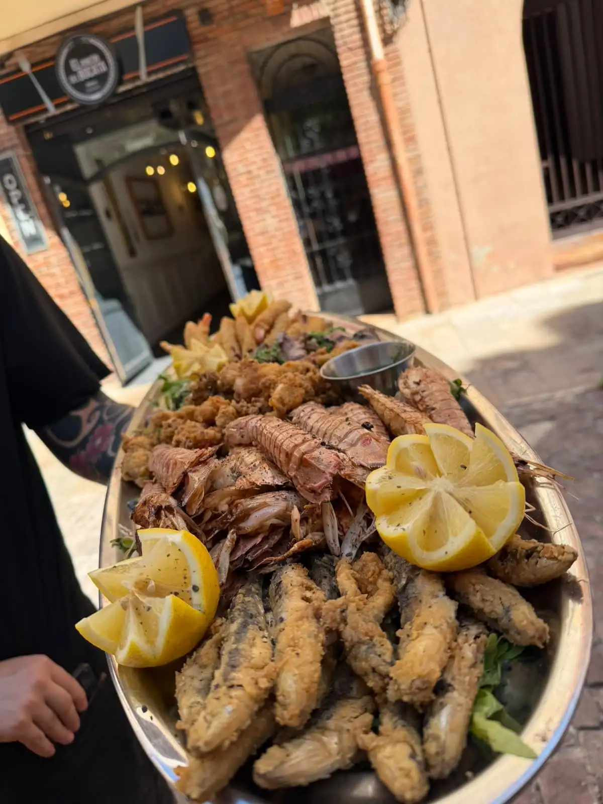 Fuente de marisco sostenida en la calle, con el rótulo y la fachada del bar al fondo: galeras, gambas, boquerones y calamares fritos con limón.