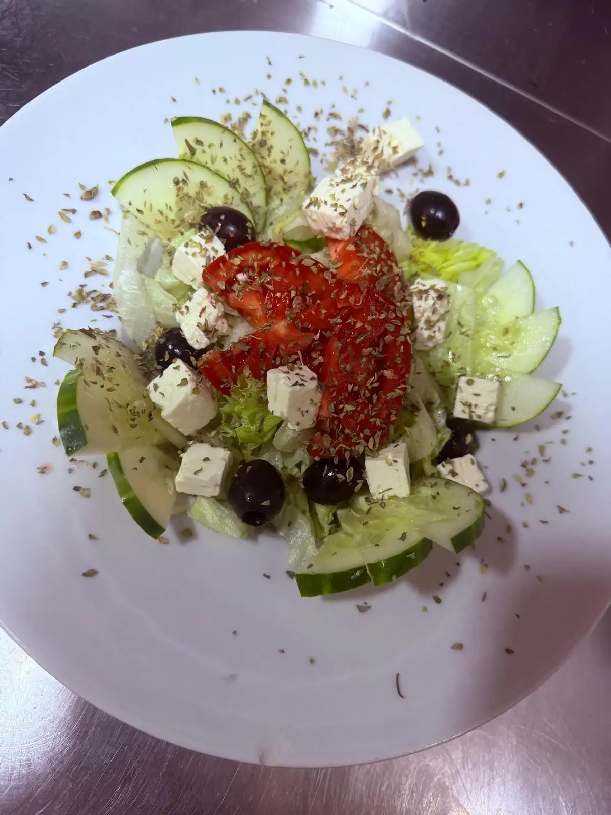 Ensalada en plato blanco: lechuga, rodajas de pepino, dados de queso feta, aceitunas negras y tomate, con orégano espolvoreado.