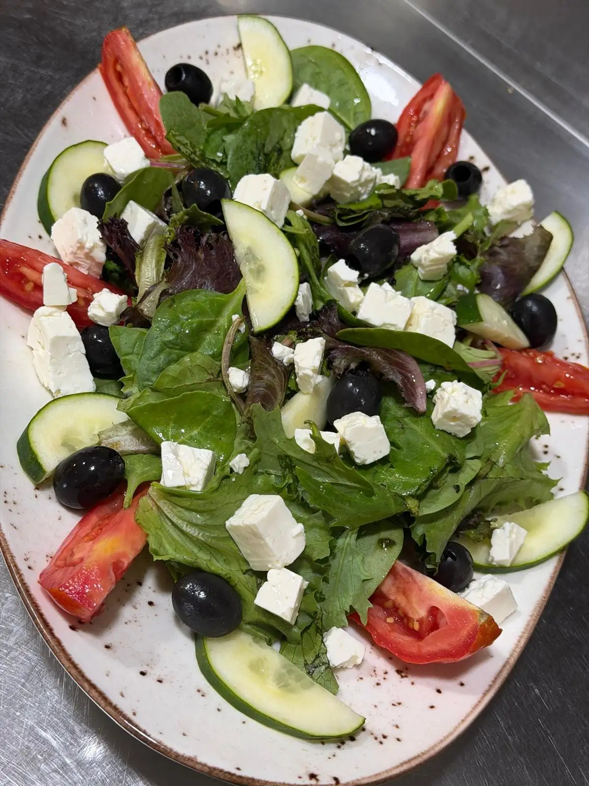 Ensalada de brotes variados en plato ovalado, con dados de queso feta, aceitunas negras, gajos de tomate y rodajas de pepino.
