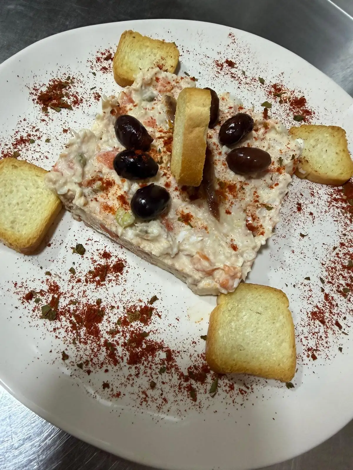 Bloque de ensaladilla cremosa moldeado en el centro del plato, con aceitunas negras y anchoas encima y tostadas alrededor, con pimentón espolvoreado.