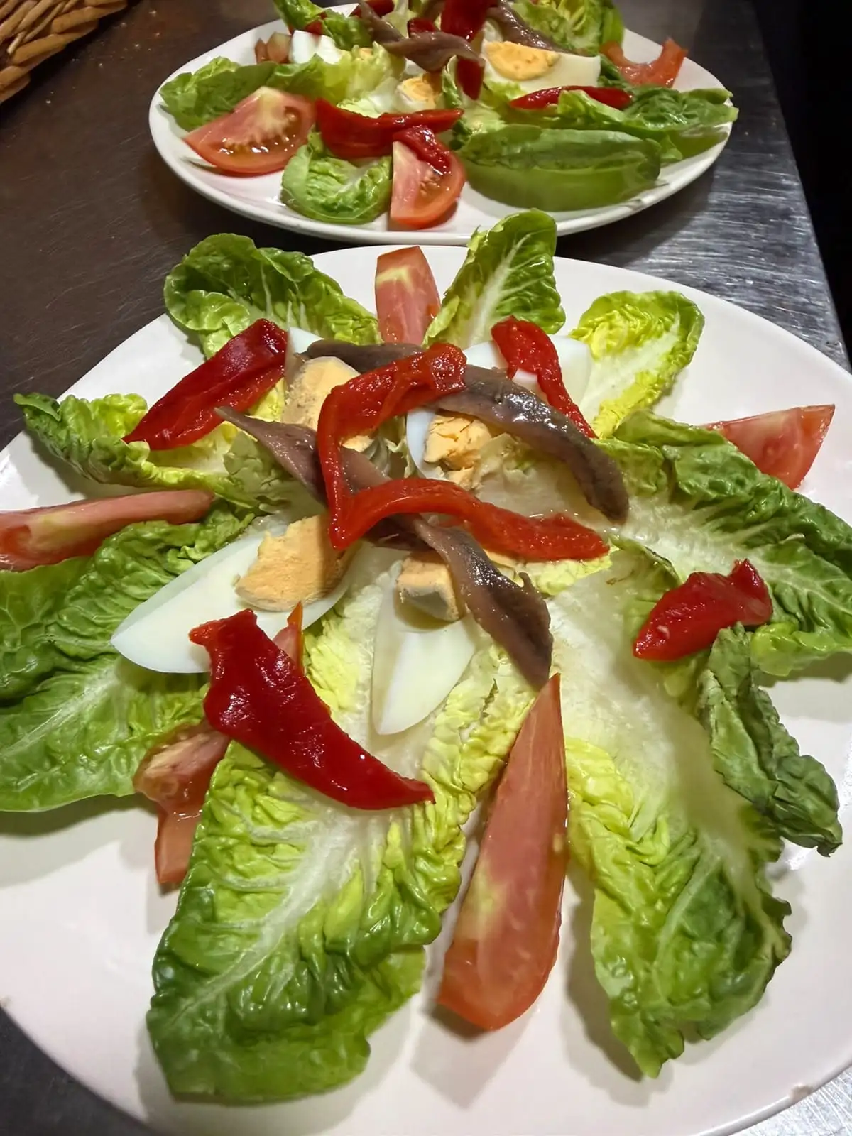 Dos platos de ensalada con hojas de cogollo, gajos de tomate, huevo duro, tiras de pimiento rojo asado y anchoas.