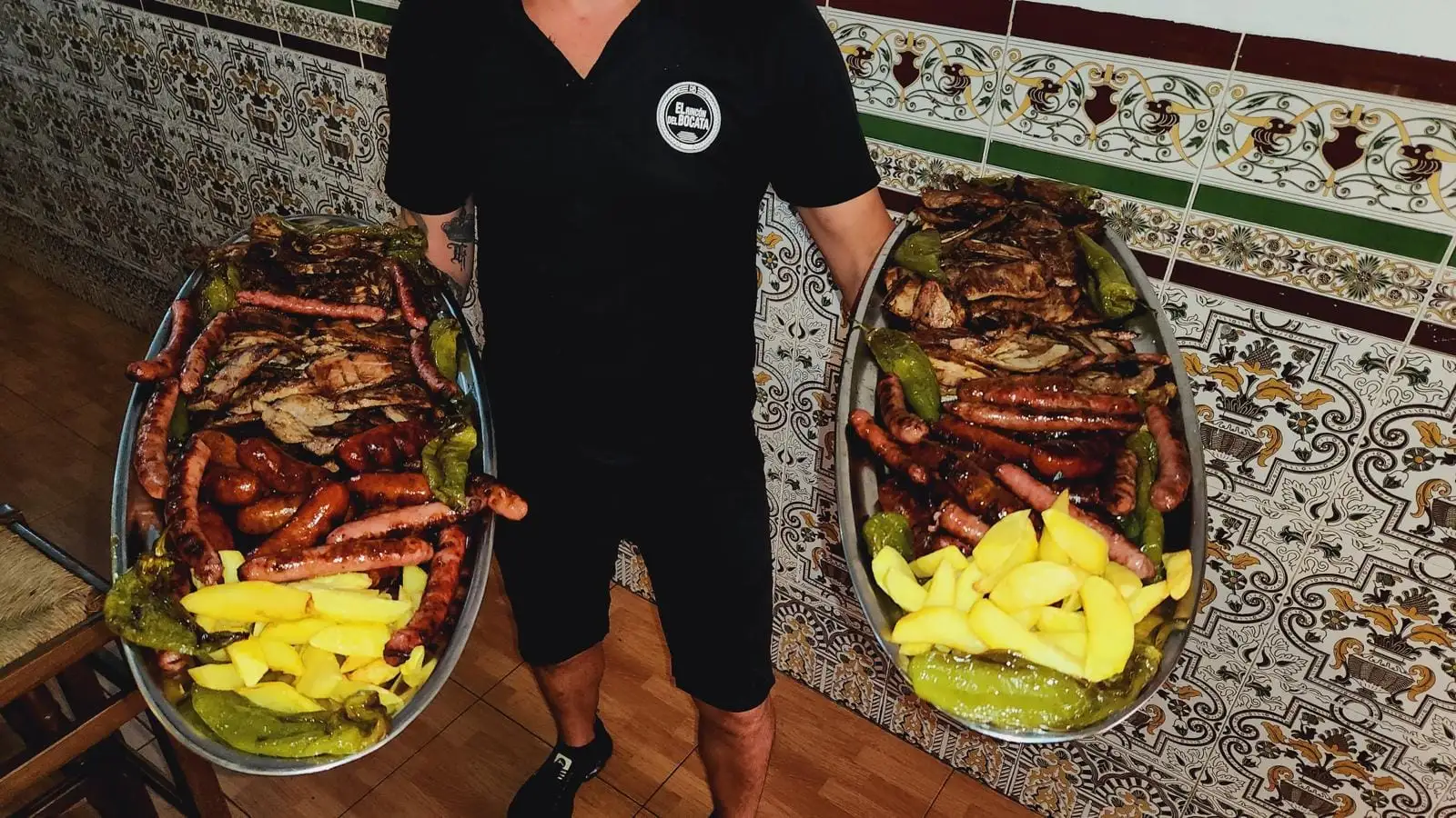 Dos grandes bandejas ovaladas de parrillada sostenidas en la sala, con longaniza, chorizo, carne a la brasa, patatas fritas y pimientos verdes.