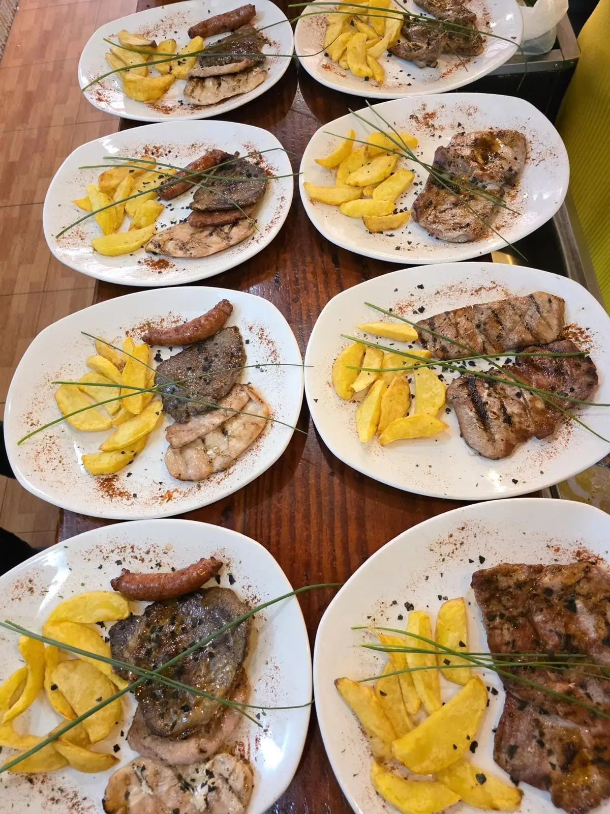 Varios platos alineados en una mesa de madera, cada uno con carne a la plancha, patatas fritas en gajos y pimentón espolvoreado en el borde.