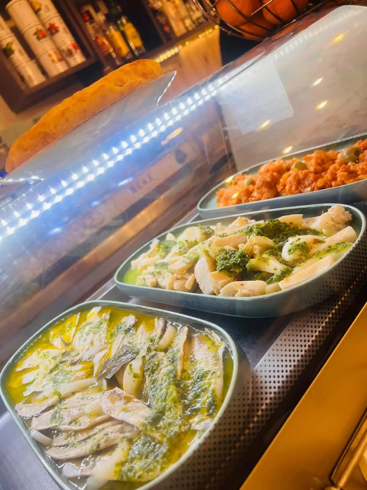 Vitrina de tapas en la barra, con bandejas de pescado en aceite y perejil, otra en salsa verde y otra con tomate y aceitunas, y un pan apoyado encima del cristal.