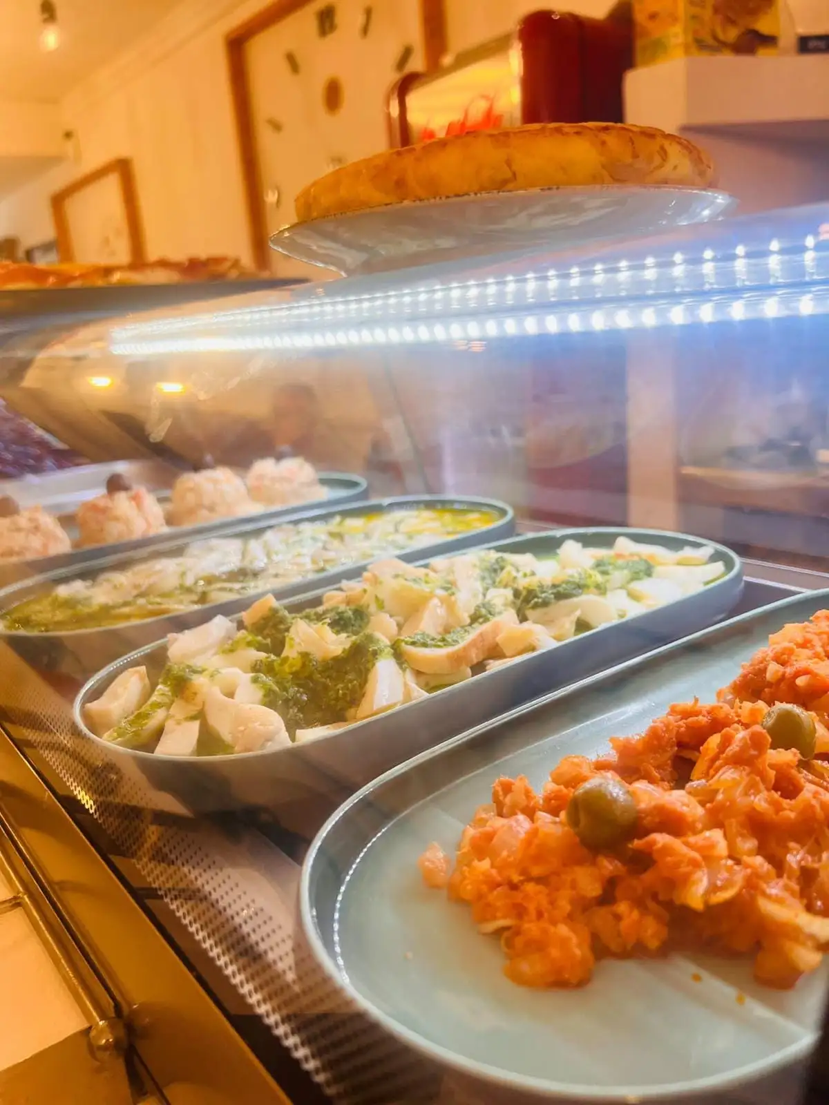 Bandejas de tapas alineadas dentro de la vitrina: pescado en aceite, trozos en salsa verde, una ensalada con tomate y aceitunas y, detrás, raciones con una gamba encima.