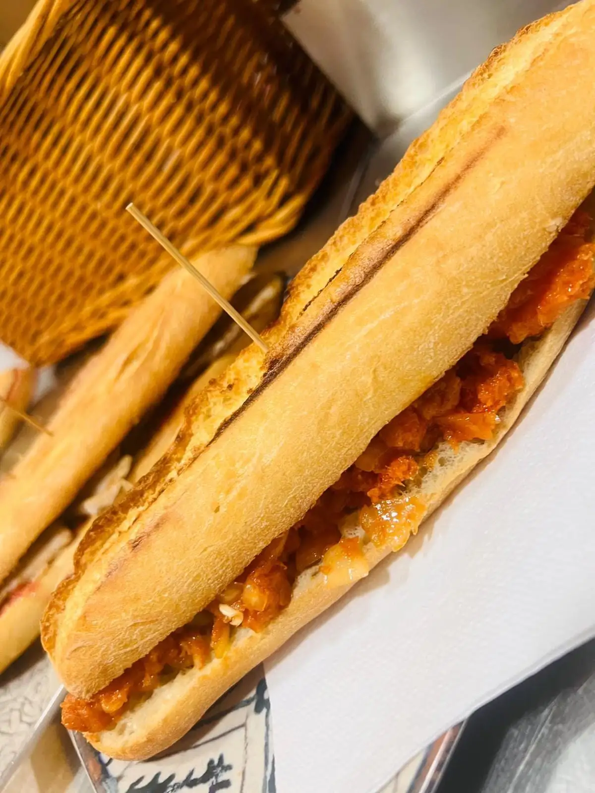 Bocadillos de pan de barra con un relleno en salsa de tomate, sujetos con palillos y servidos sobre papel junto a un cesto de mimbre.