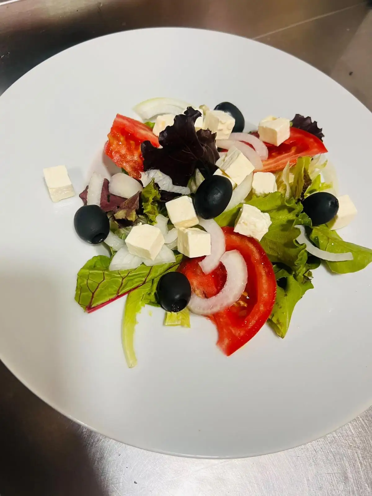 Ensalada de brotes variados con gajos de tomate, cebolla en aros, aceitunas negras y dados de queso blanco, en un plato blanco.