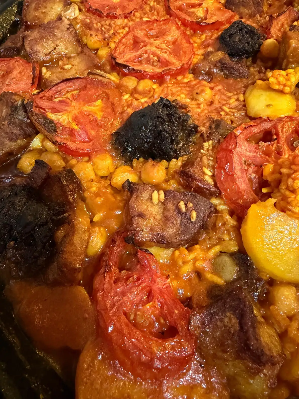Primer plano de un arroz al horno en la cazuela: rodajas de tomate asado, trozos de morcilla, garbanzos, tacos de carne y rodajas de patata entre el grano tostado.