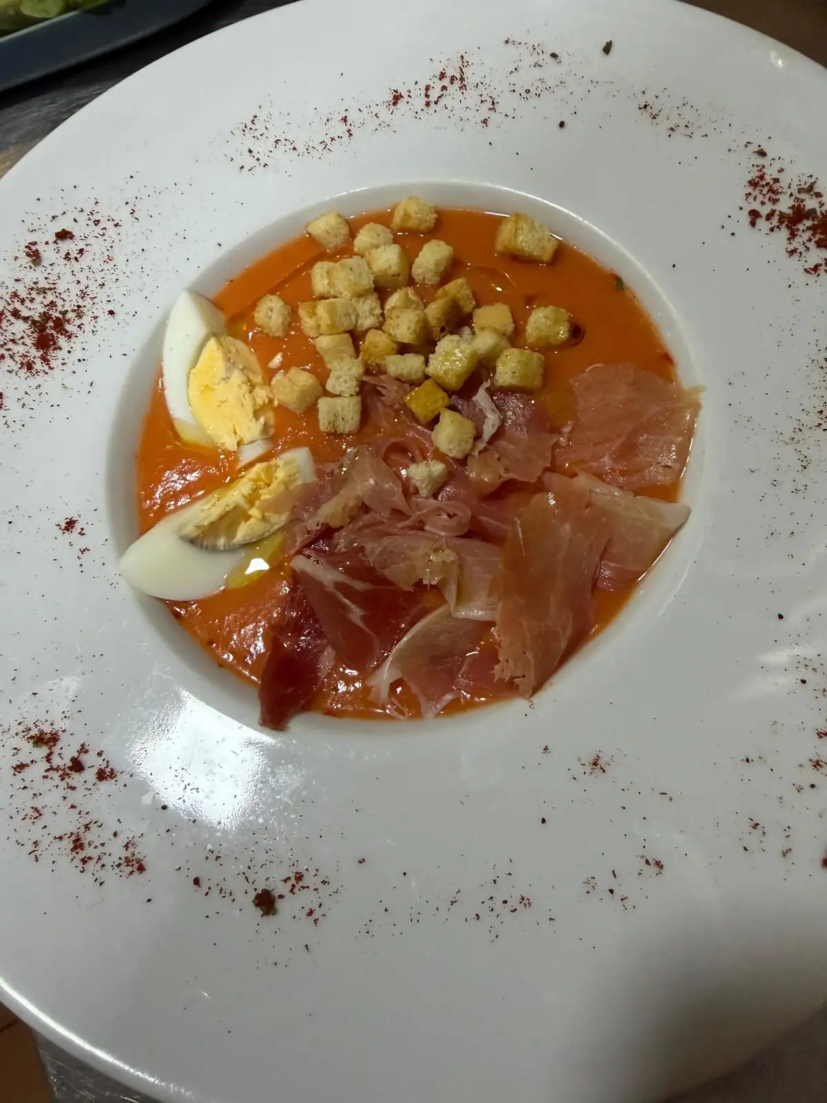 Plato hondo blanco con crema fría de tomate, cuartos de huevo duro, picatostes y lonchas de jamón serrano, con pimentón espolvoreado por todo el borde.