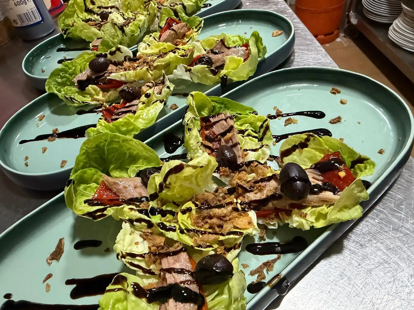 Hilera de platos ovalados de loza verde con hojas de cogollo rellenas de atún, tomate y aceituna negra, con cebolla frita crujiente y trazos de reducción de balsámico.