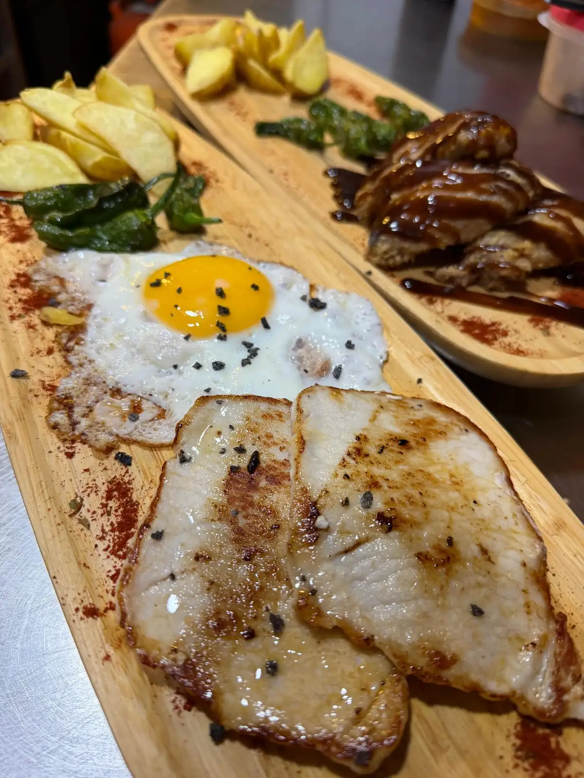 Tabla alargada de madera con dos filetes de lomo a la plancha, huevo frito, patatas fritas y pimientos de Padrón; detrás, otra tabla con carne bañada en salsa oscura y patatas en gajos.
