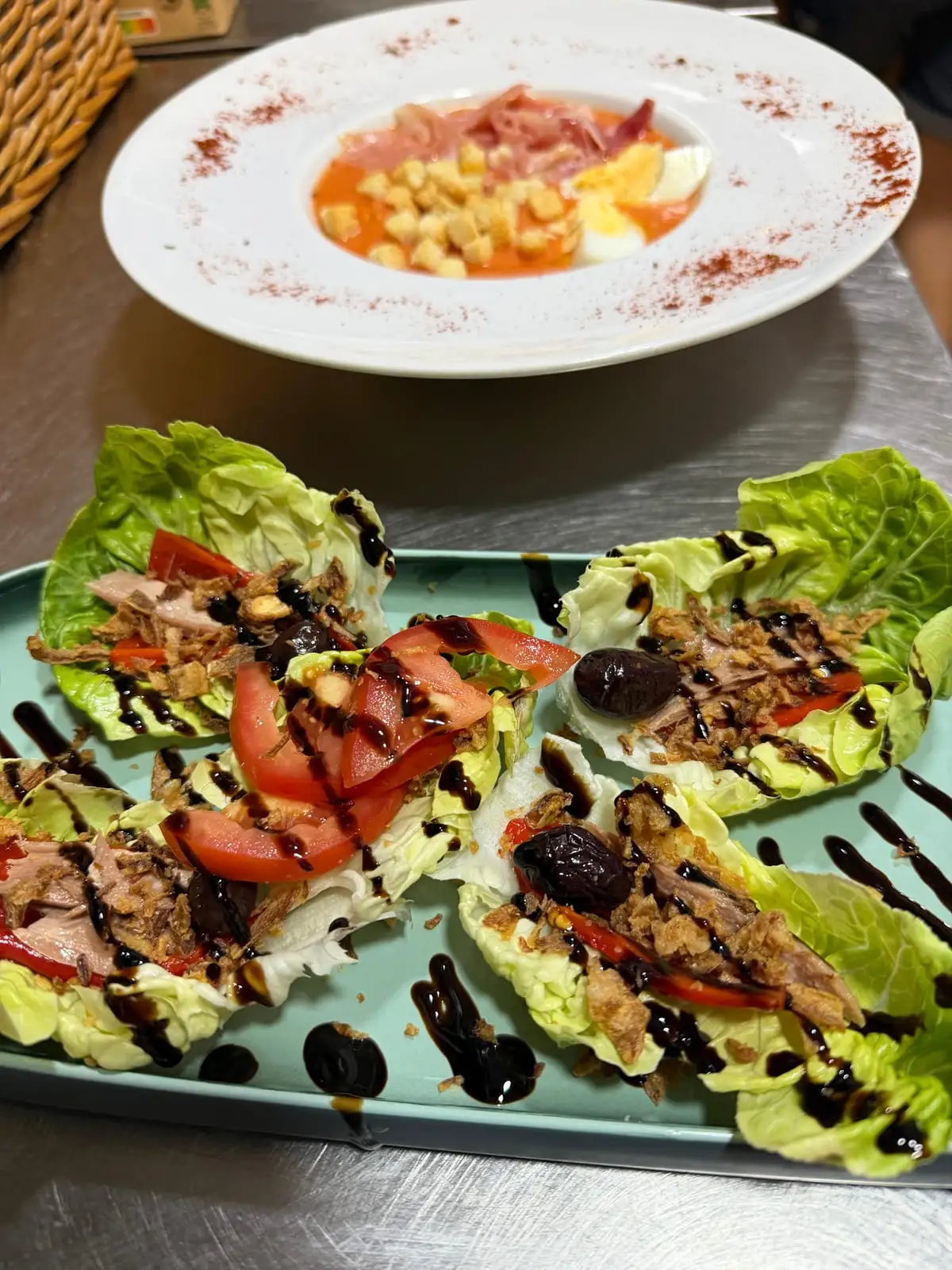 Bandeja verde con cuatro hojas de cogollo rellenas de atún, tomate y aceituna negra con reducción de balsámico, y al fondo un plato de crema fría de tomate con huevo duro, picatostes y jamón.