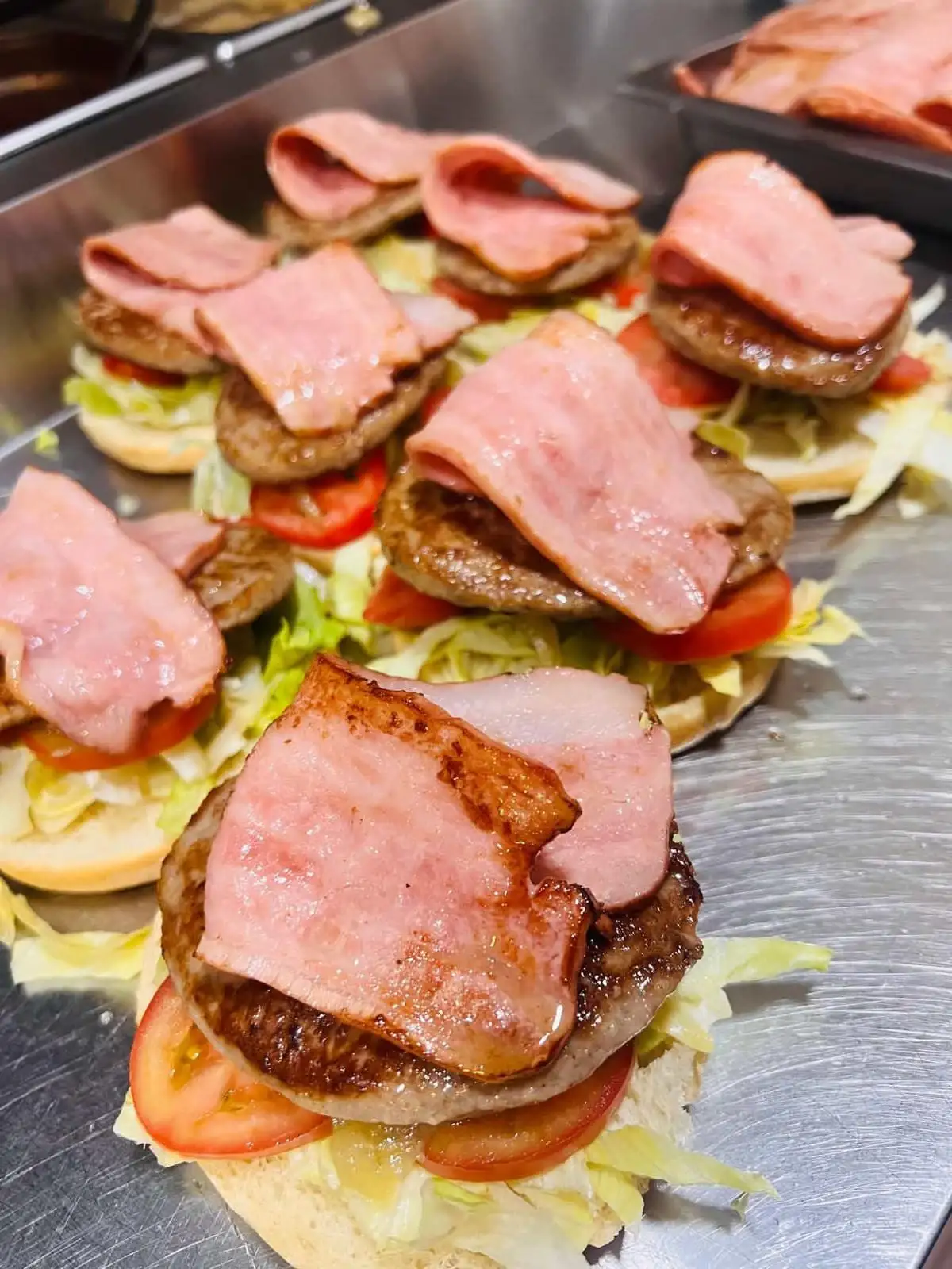 Panes redondos abiertos en fila sobre la plancha, cada uno con lechuga, rodajas de tomate, una hamburguesa a la plancha y lonchas de bacon por encima.
