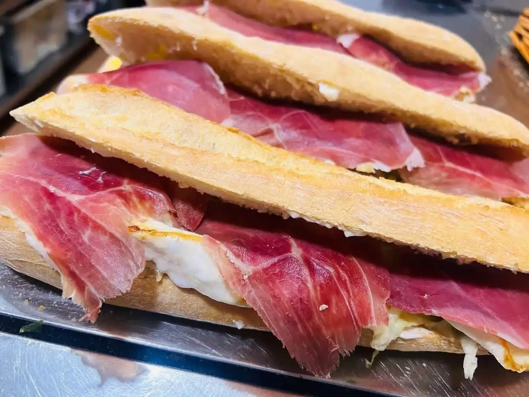 Bocadillos de barra alineados en una bandeja, abiertos y cubiertos de lonchas finas de jamón serrano.