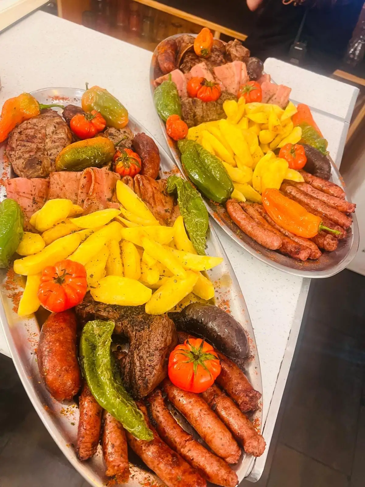 Dos bandejas ovaladas de parrillada servidas en la sala: filetes y carne a la brasa, morcilla, longanizas, patatas fritas en gajos, pimientos verdes y tomate asado.