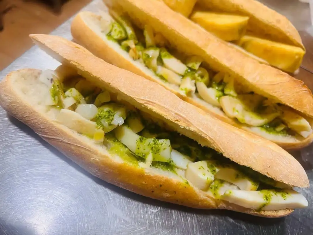 Dos bocadillos de barra abiertos, rellenos de sepia troceada en salsa verde de ajo y perejil, con patatas fritas al fondo.