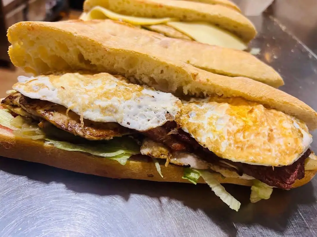 Bocadillo de chapata con lechuga, lomo a la plancha, una tira de embutido rojo y huevo frito, con otro de queso al fondo.