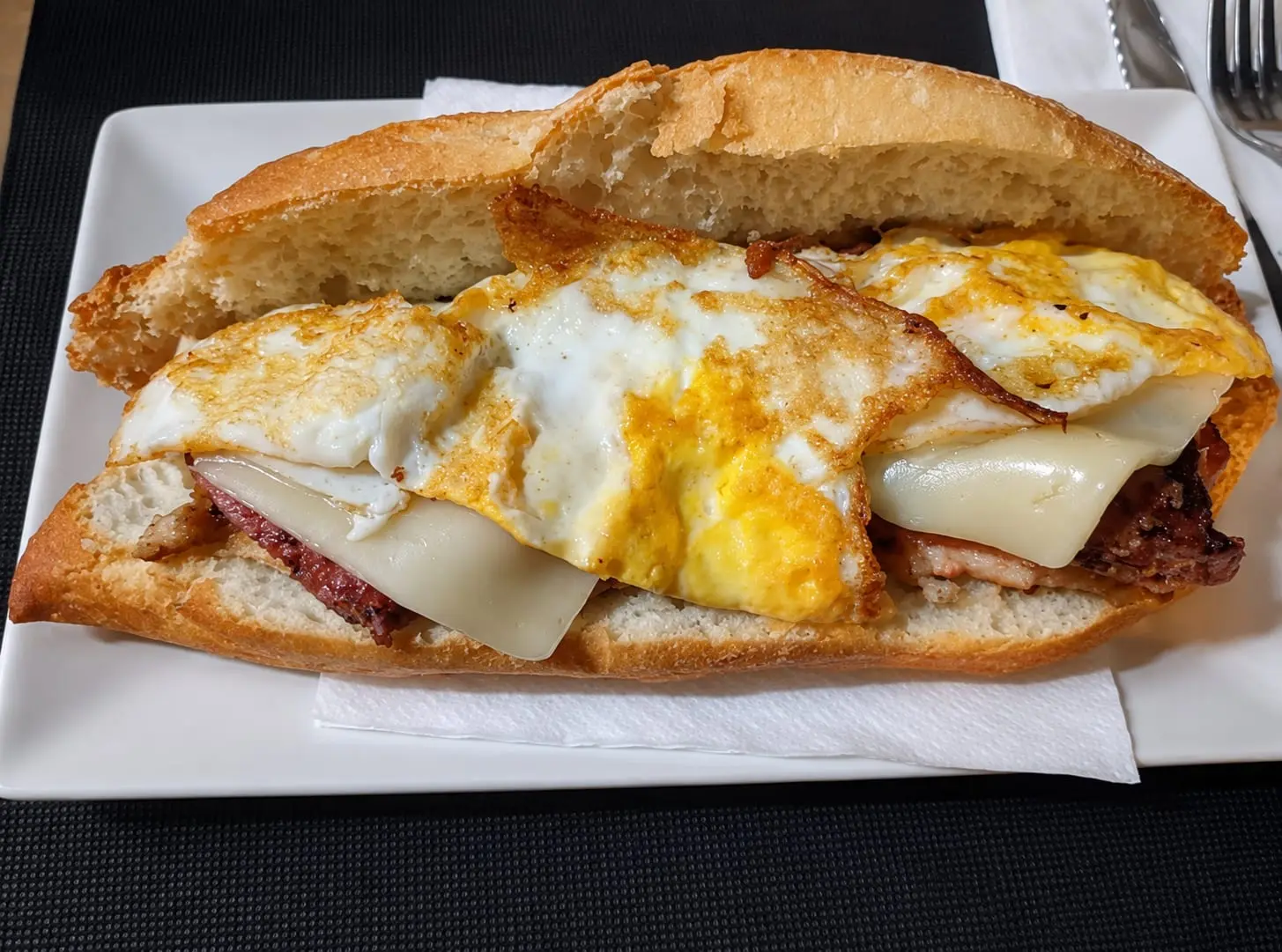 Bocadillo abierto en un plato blanco, con carne a la plancha, queso fundido y huevo frito encima.