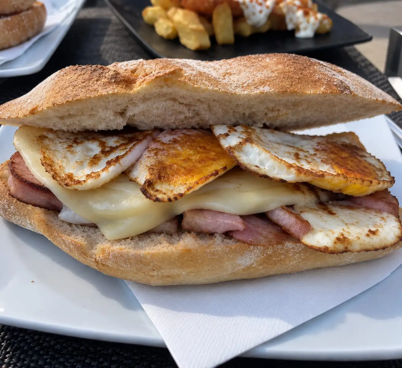 Bocadillo de pan crujiente con lomo, queso fundido y dos huevos fritos, servido en la terraza.