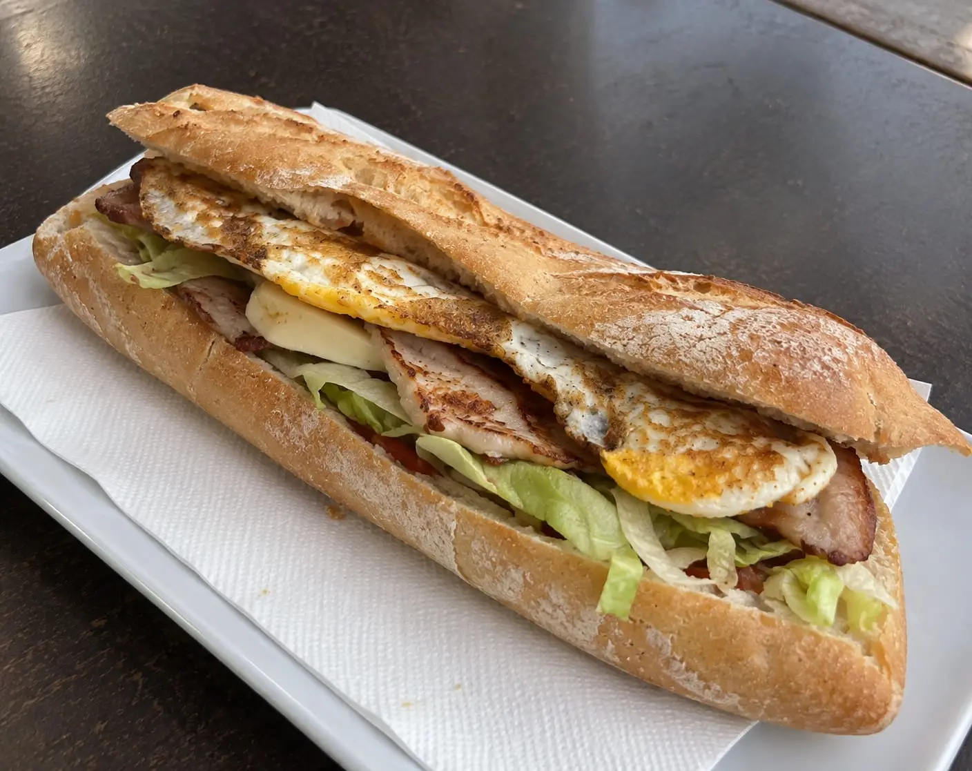 Bocadillo de barra larga con lomo, queso, lechuga y huevo frito, servido en un plato blanco.