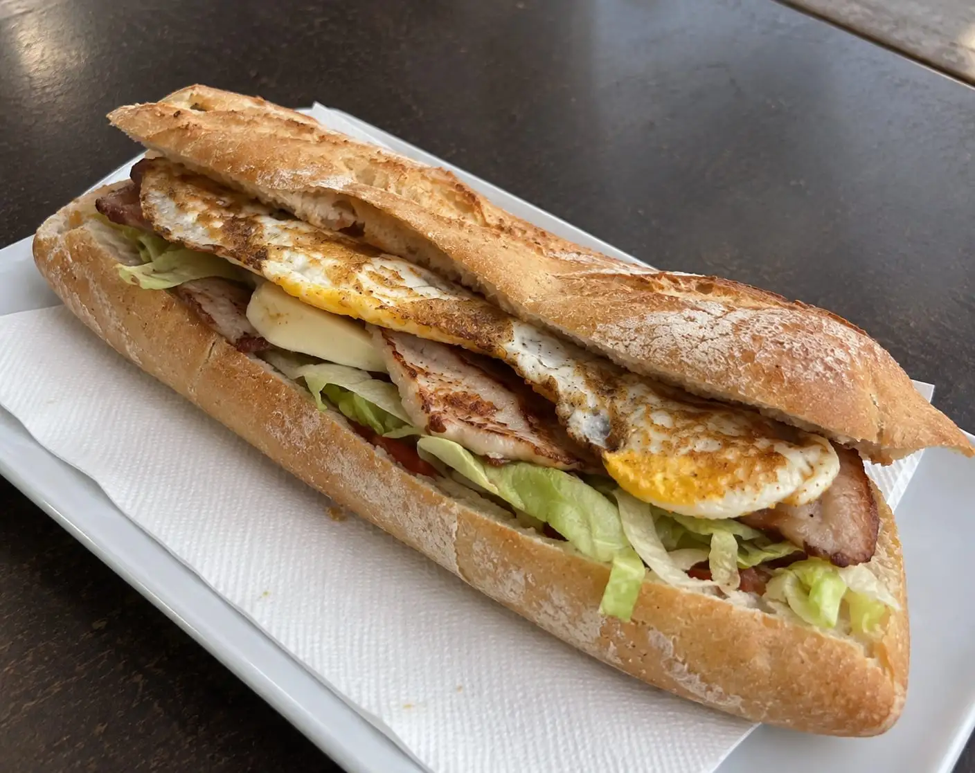 Bocadillo de barra larga con lomo, queso, lechuga y huevo frito, servido en un plato blanco.