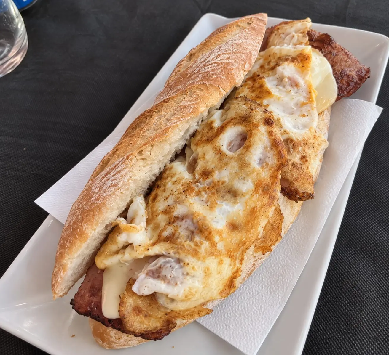 Bocadillo abierto con tortilla, bacon y queso fundido, servido en un plato blanco.