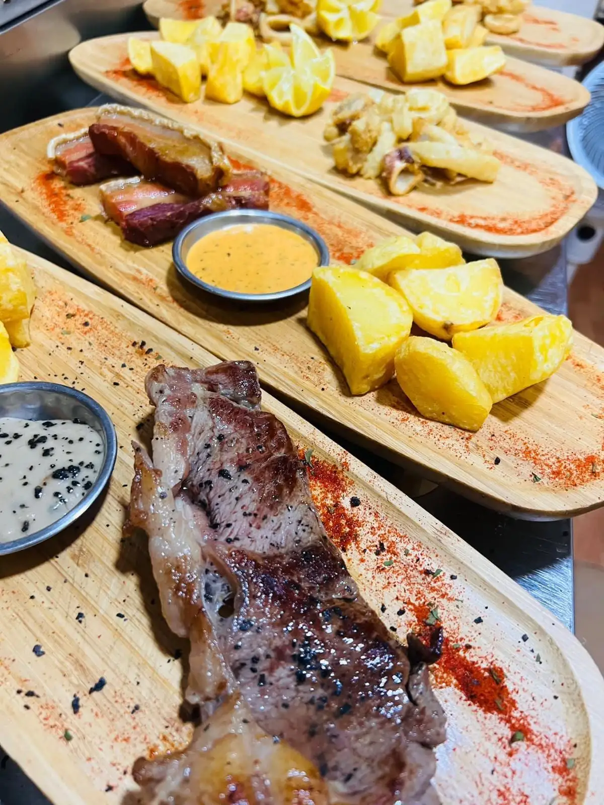 Hilera de tablas de madera recién servidas: entrecot, panceta, calamares, patatas y limón tallado.