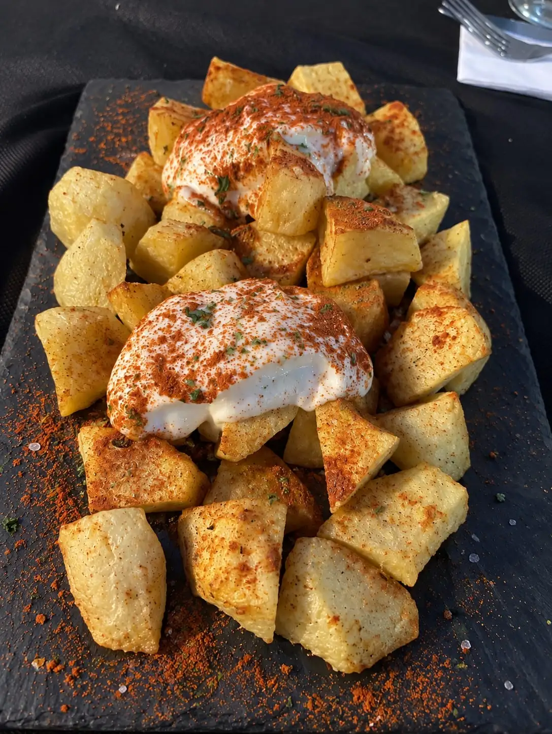 Patatas bravas servidas en una pizarra, con dos montones de salsa blanca espolvoreada de pimentón.