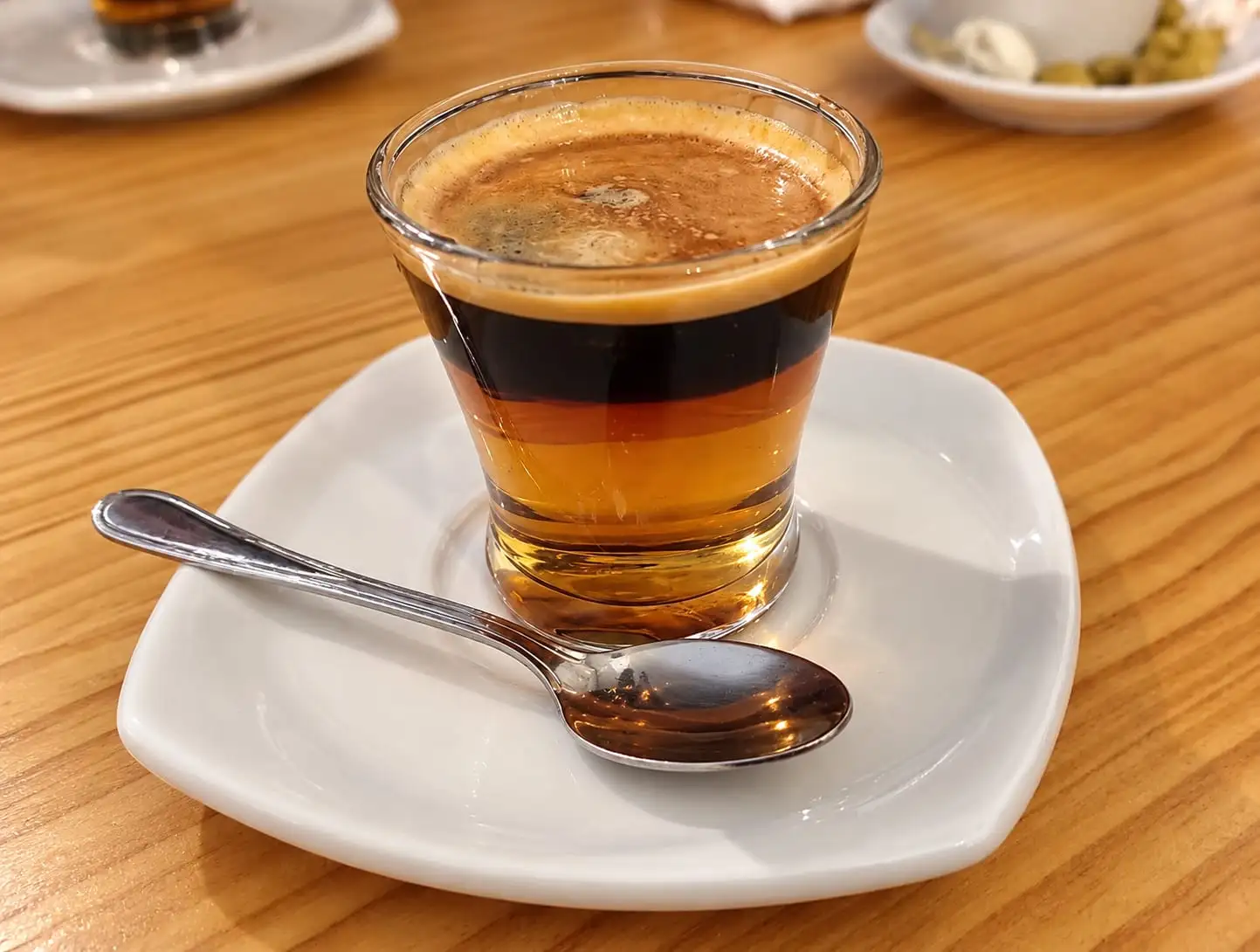 Café en vaso de cristal con las capas a la vista -leche condensada, café y crema- sobre un platillo blanco.