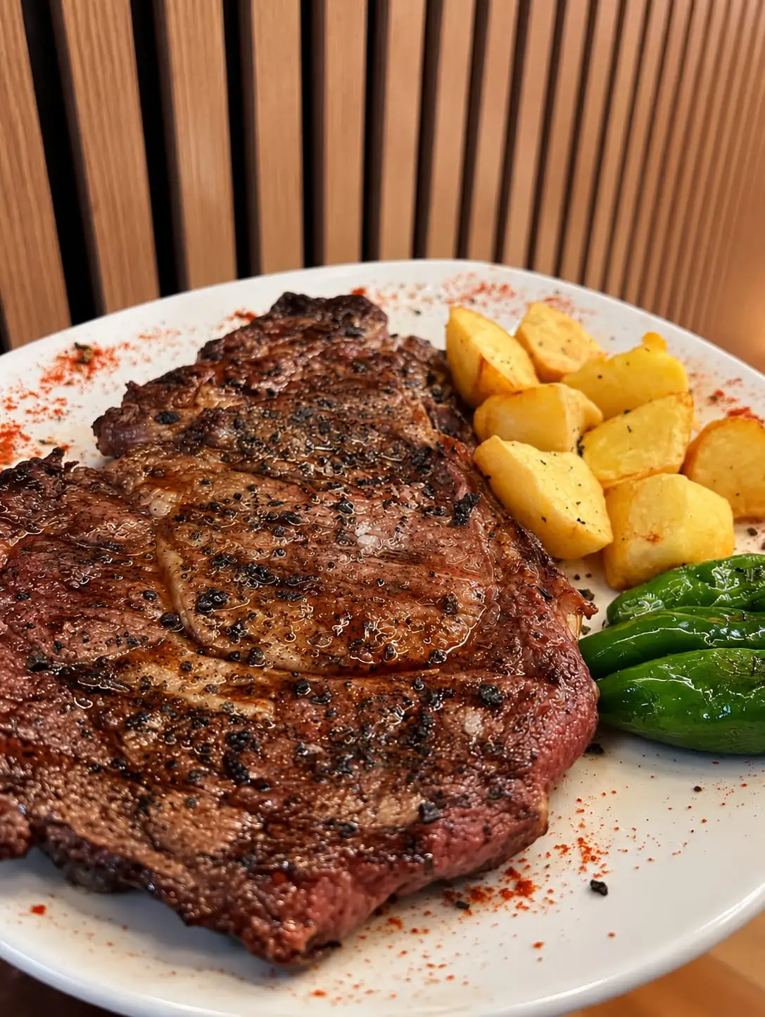 Chuletón hecho a la brasa en un plato blanco, con patatas y pimientos verdes al lado.