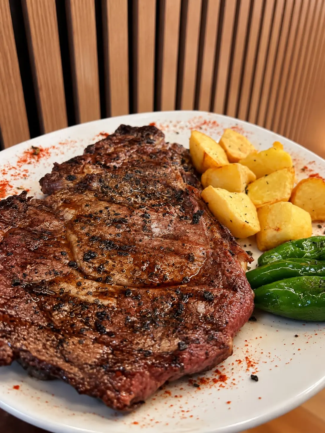Chuletón hecho a la brasa en un plato blanco, con patatas y pimientos verdes al lado.